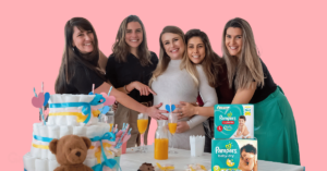 a pregnant woman surrounded by her friends during a baby shower with two packs of pampers diapers on the table in front of them.